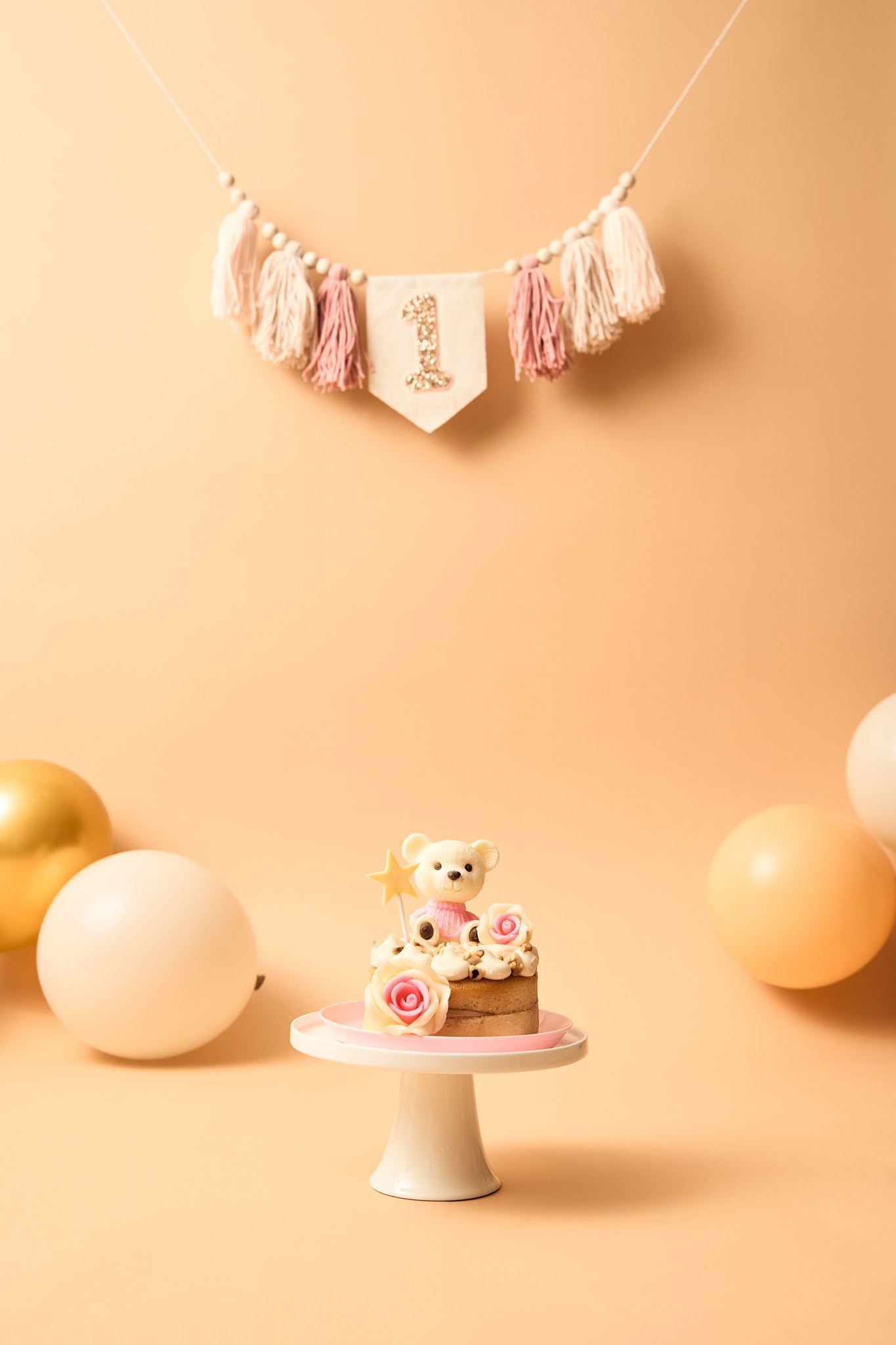 A small single-tier smash cake with pale buttercream icing and a bear topper on a white cake stand, styled against a peach backdrop
