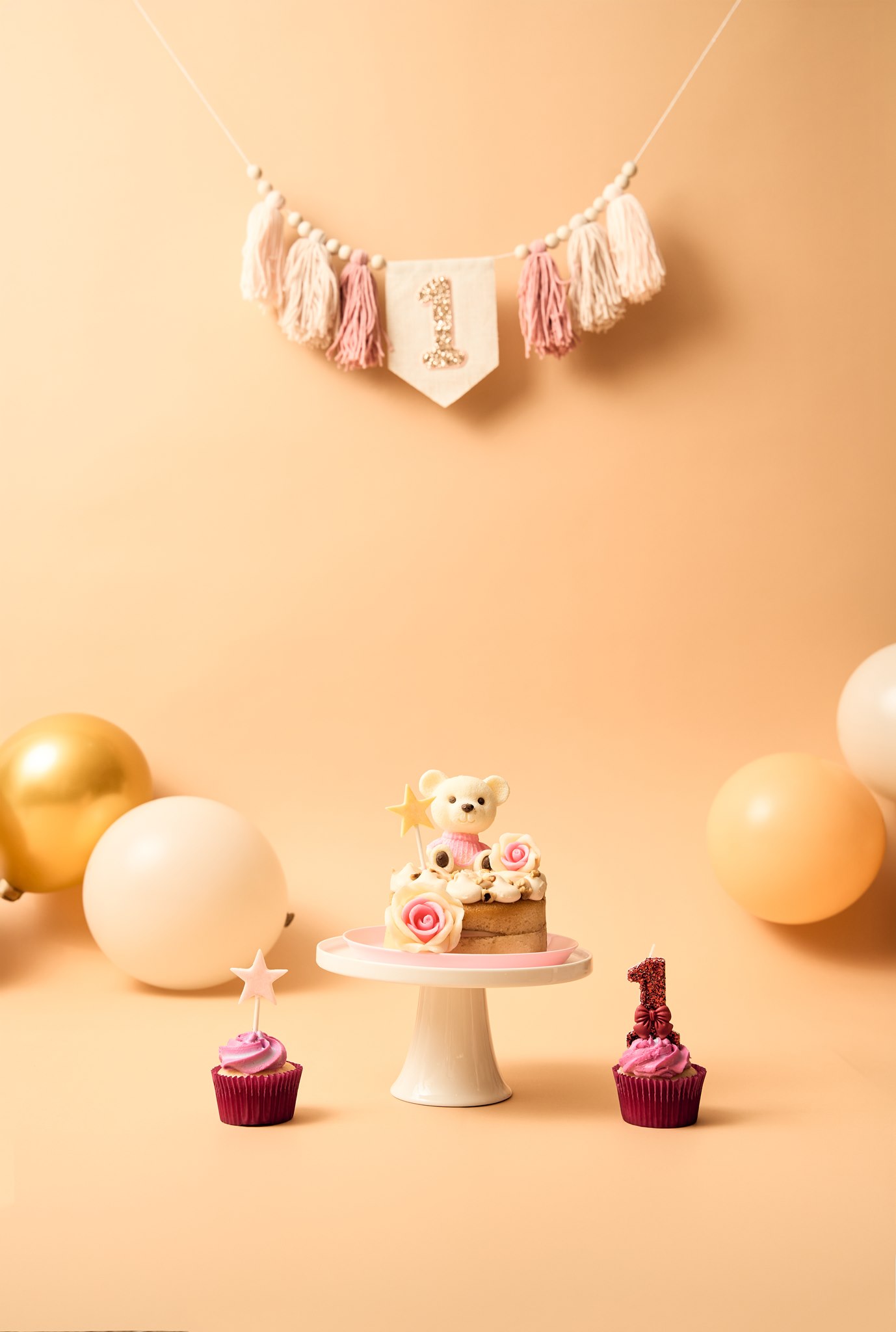 A styled cake smash setup with peach backdrop, balloons, a number one banner and the smash cake on a stand, ready in a family's home