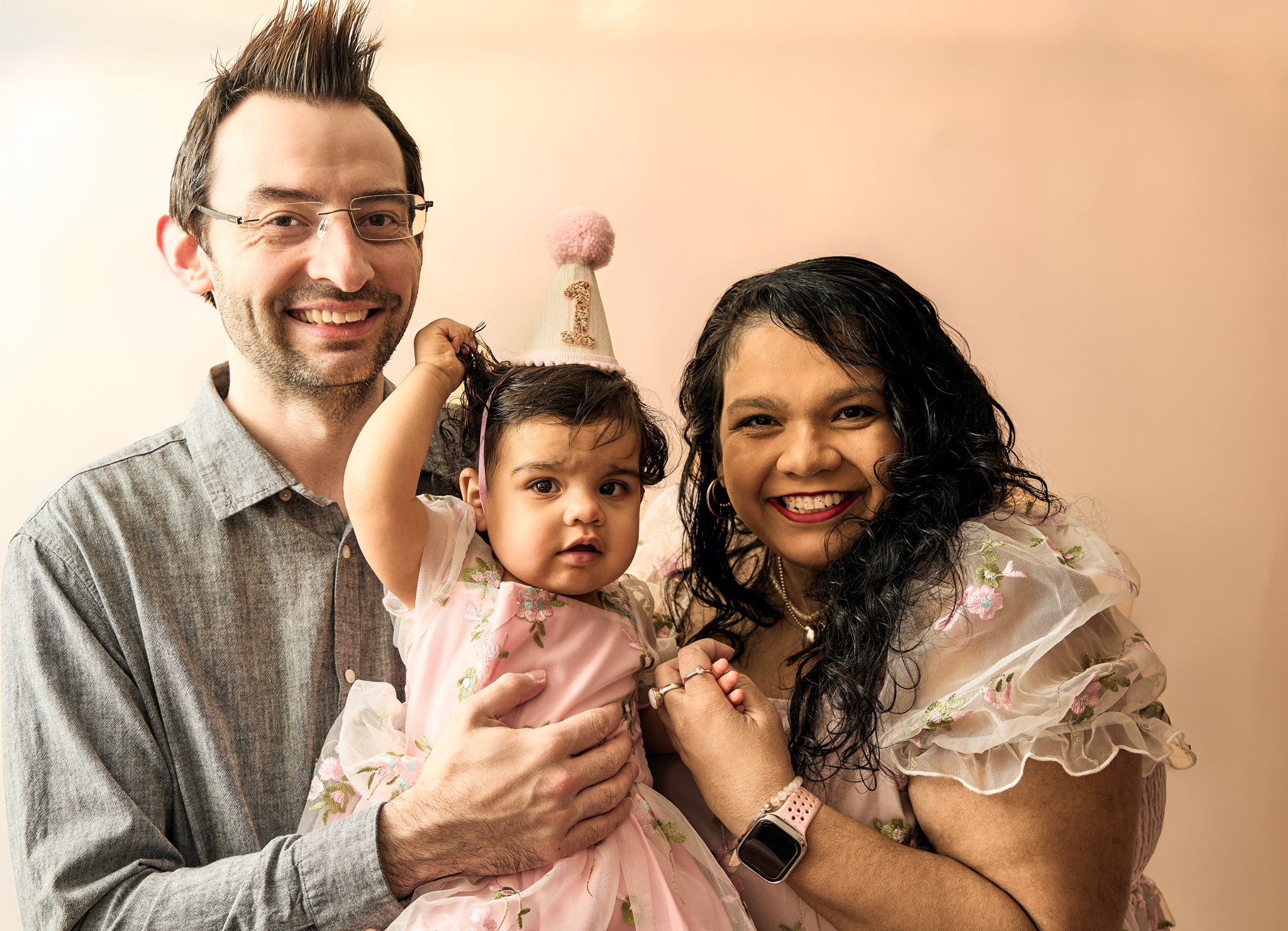 Mum and dad holding their one-year-old daughter in a party hat, smiling together before the cake smash begins