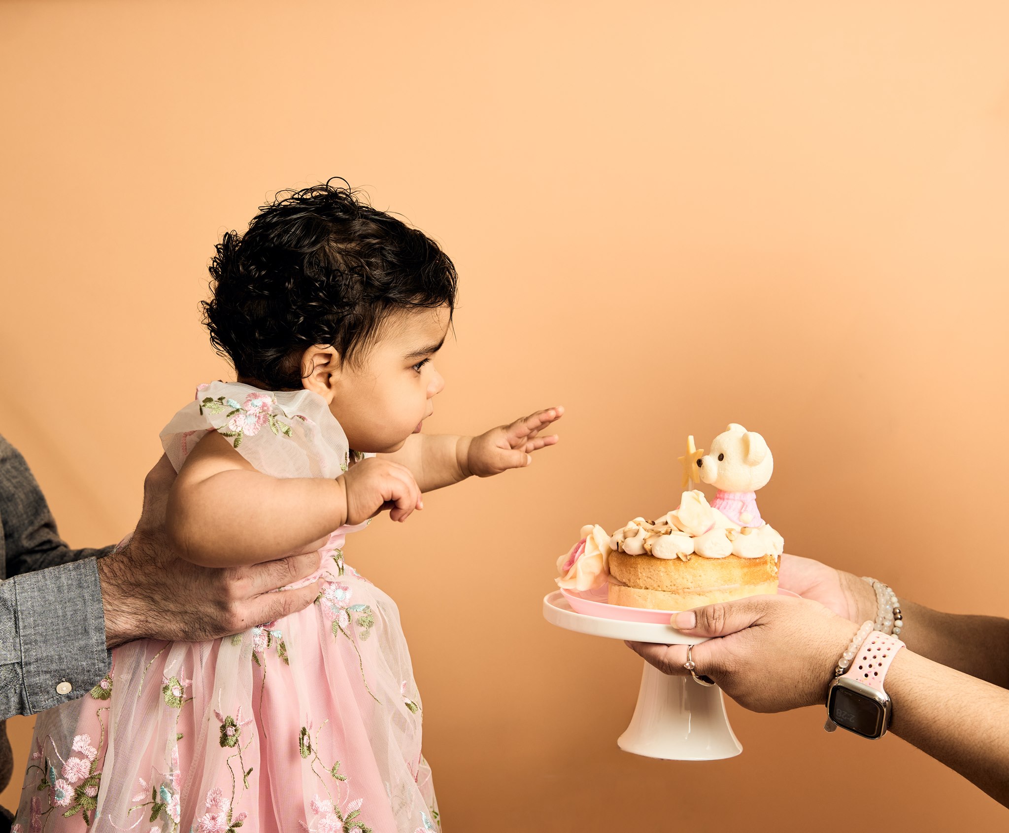 A one-year-old reaching cautiously towards her buttercream smash cake as it is held out to her during a first birthday session