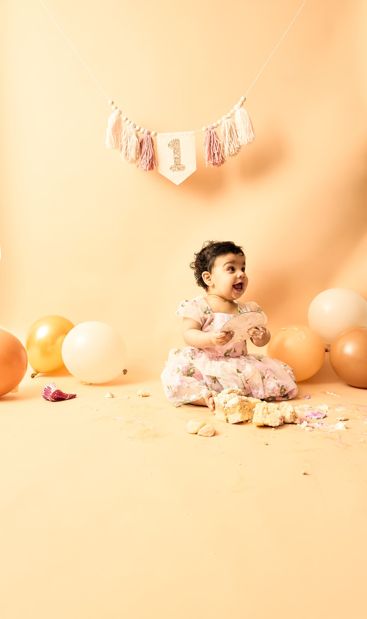 Wide photo of the aftermath of a cake smash session, crumbs and icing spread across the floor around a satisfied one-year-old