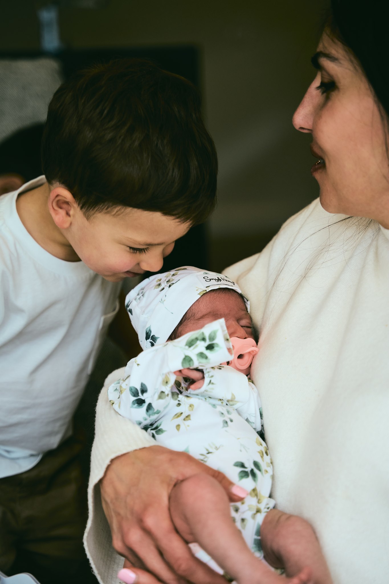Big brother gazing down at his newborn sibling swaddled in mum's arms, in-home family session