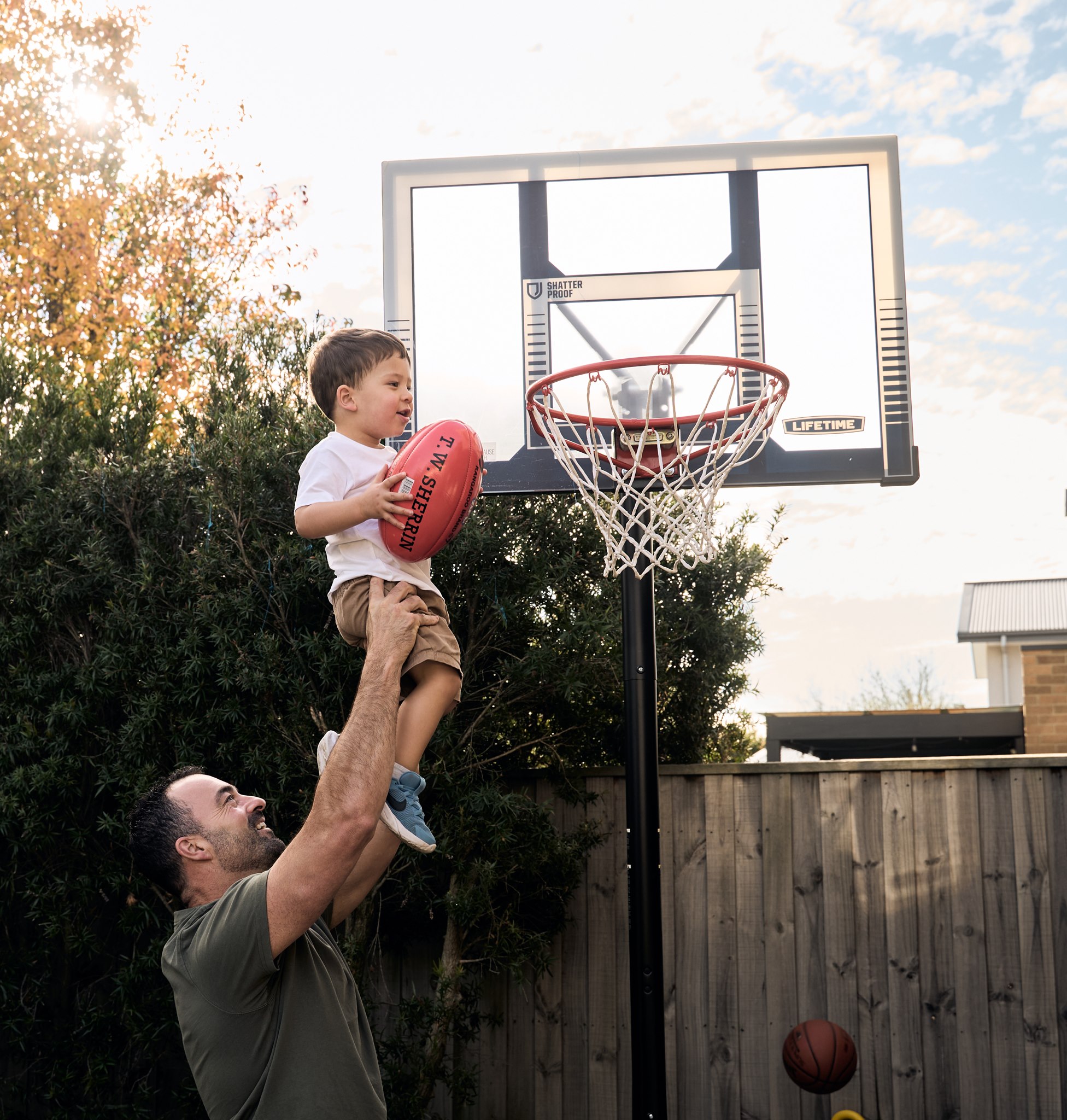 Dad lifting his laughing son into the air by the basketball hoop, outdoor lifestyle family session