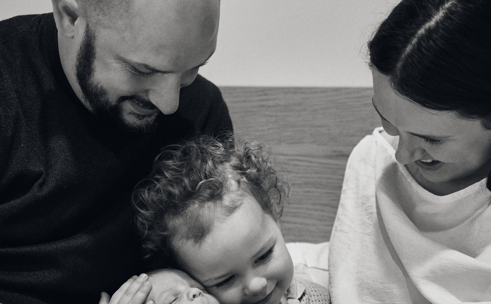 Black and white photo of a toddler leaning in to kiss the new baby in mum's arms