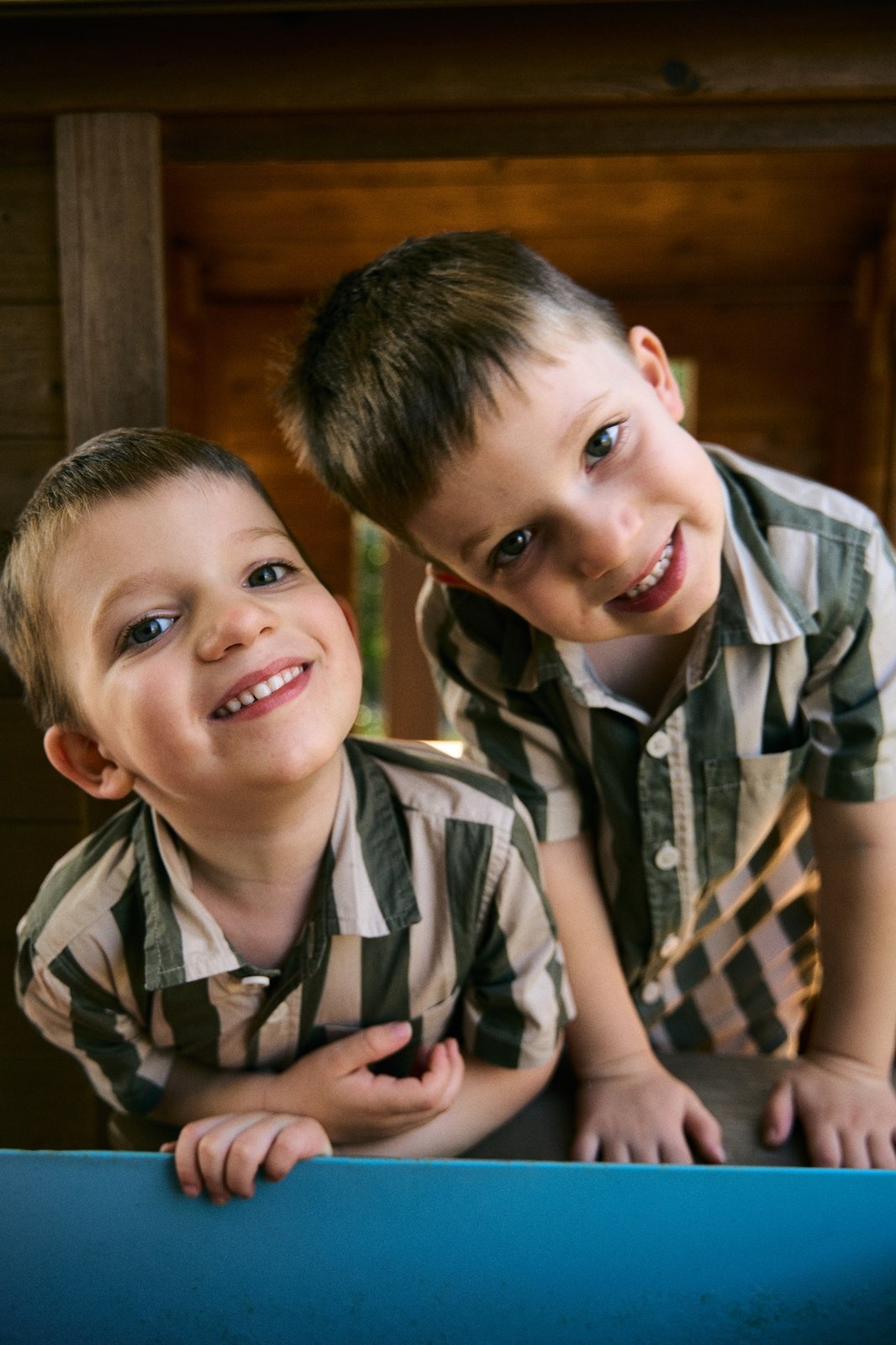 Two young brothers grinning side by side at their cubby house