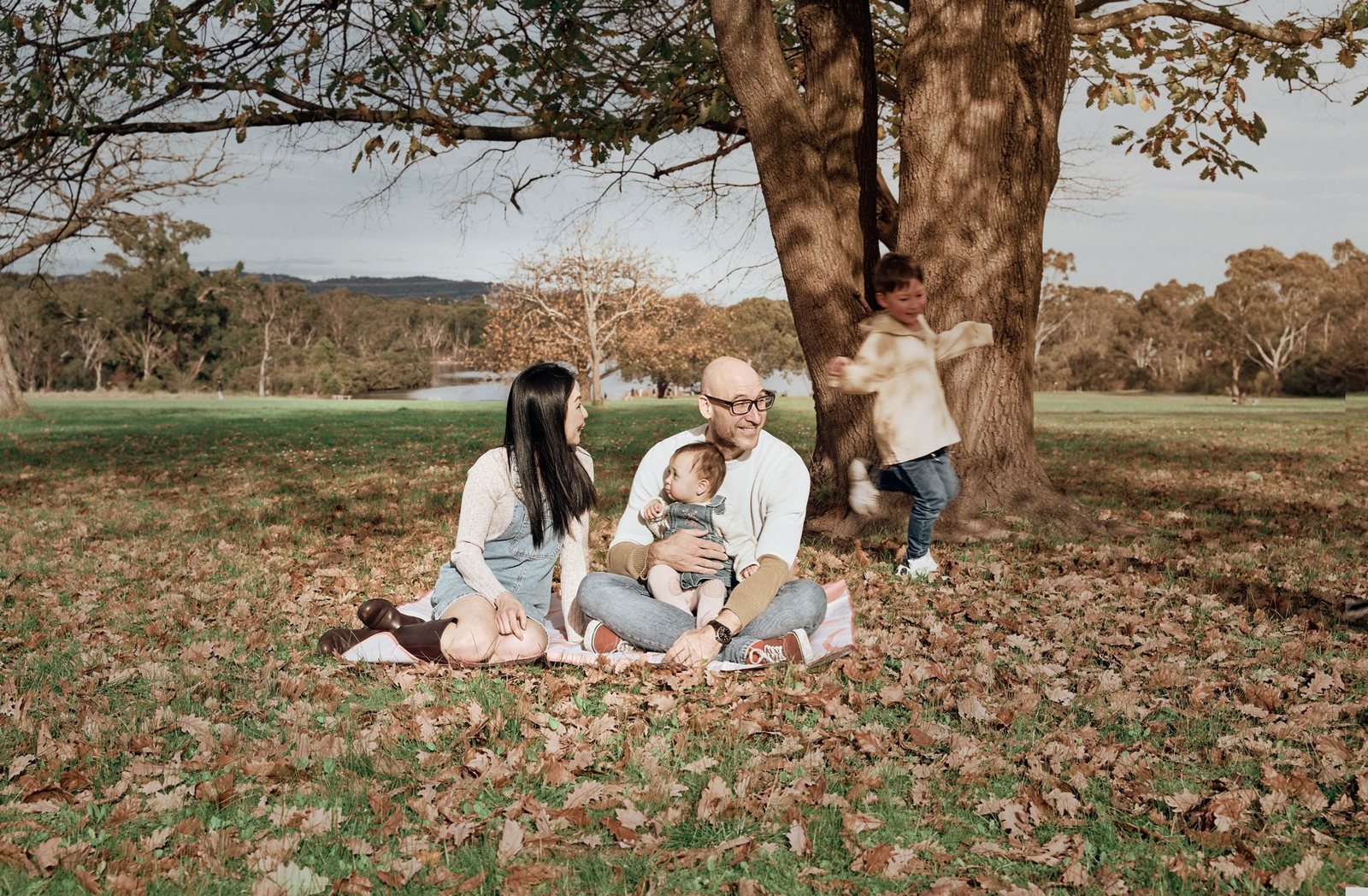 Family sitting on a rug under a tree while their toddler runs past