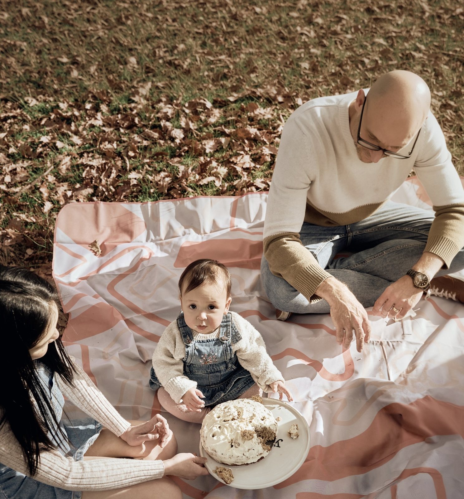 Parents watching their baby with a smash cake on a picnic rug in autumn leaves