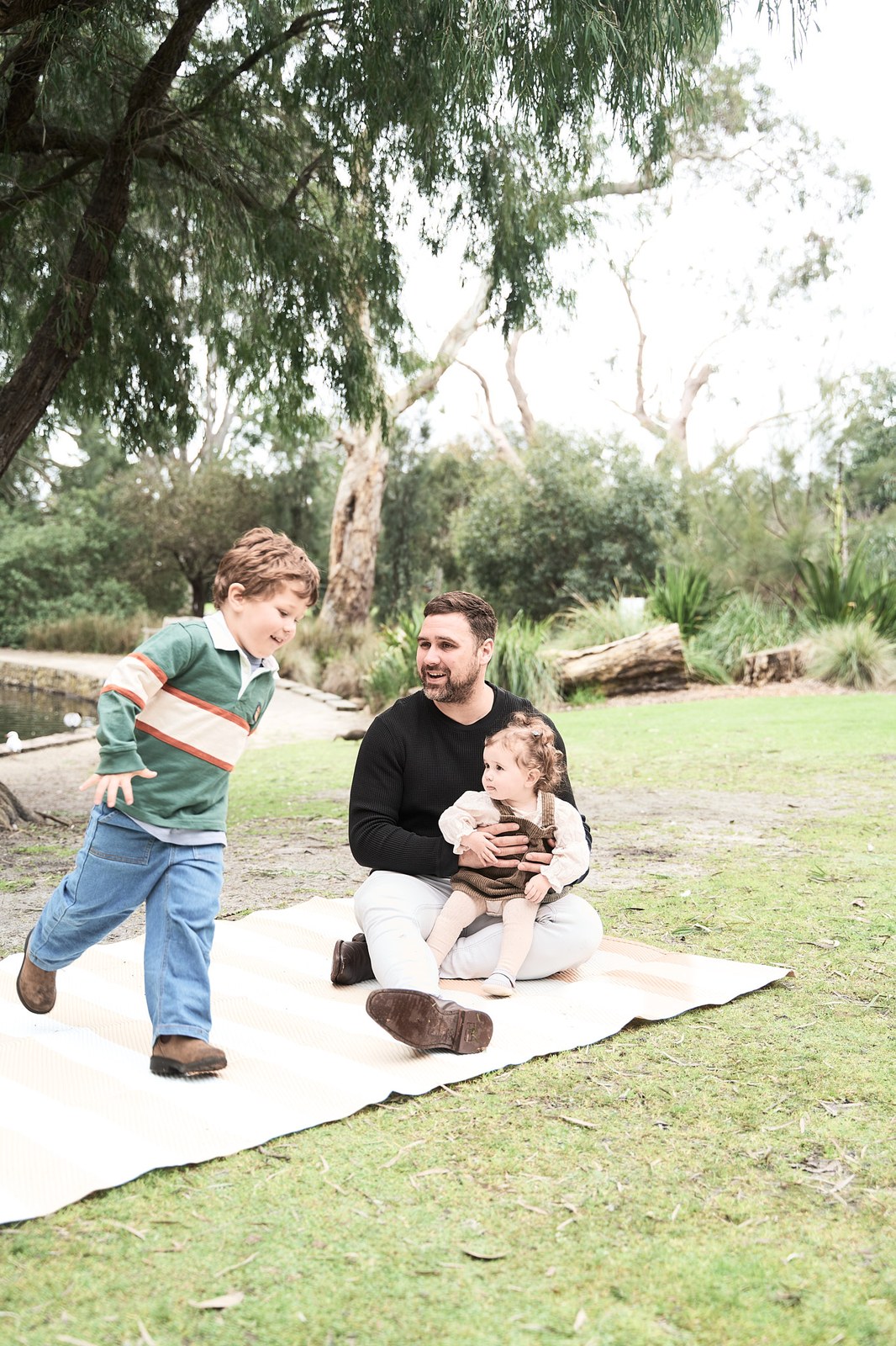 Little boy running past his dad and baby sister on the picnic rug