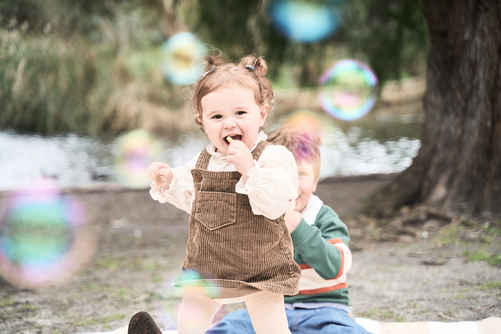 Toddler in a cord pinafore playing with bubbles by the water