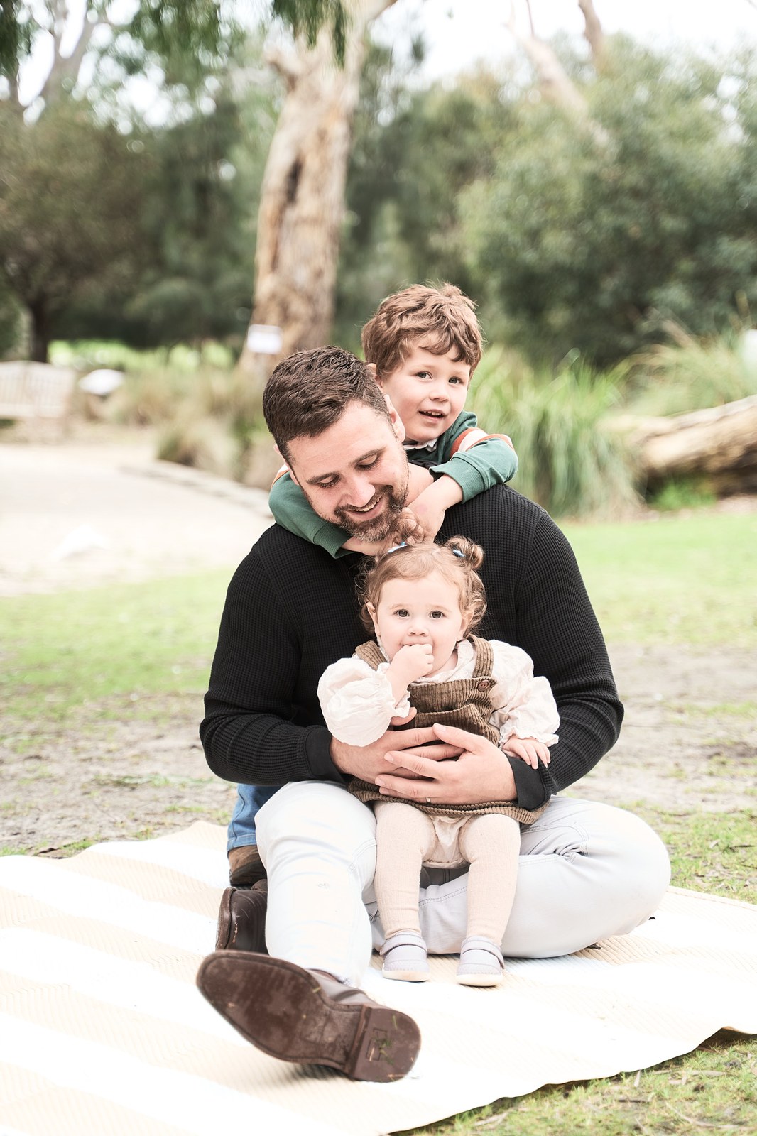 Dad on a picnic rug with his son on his shoulders and his baby on his lap