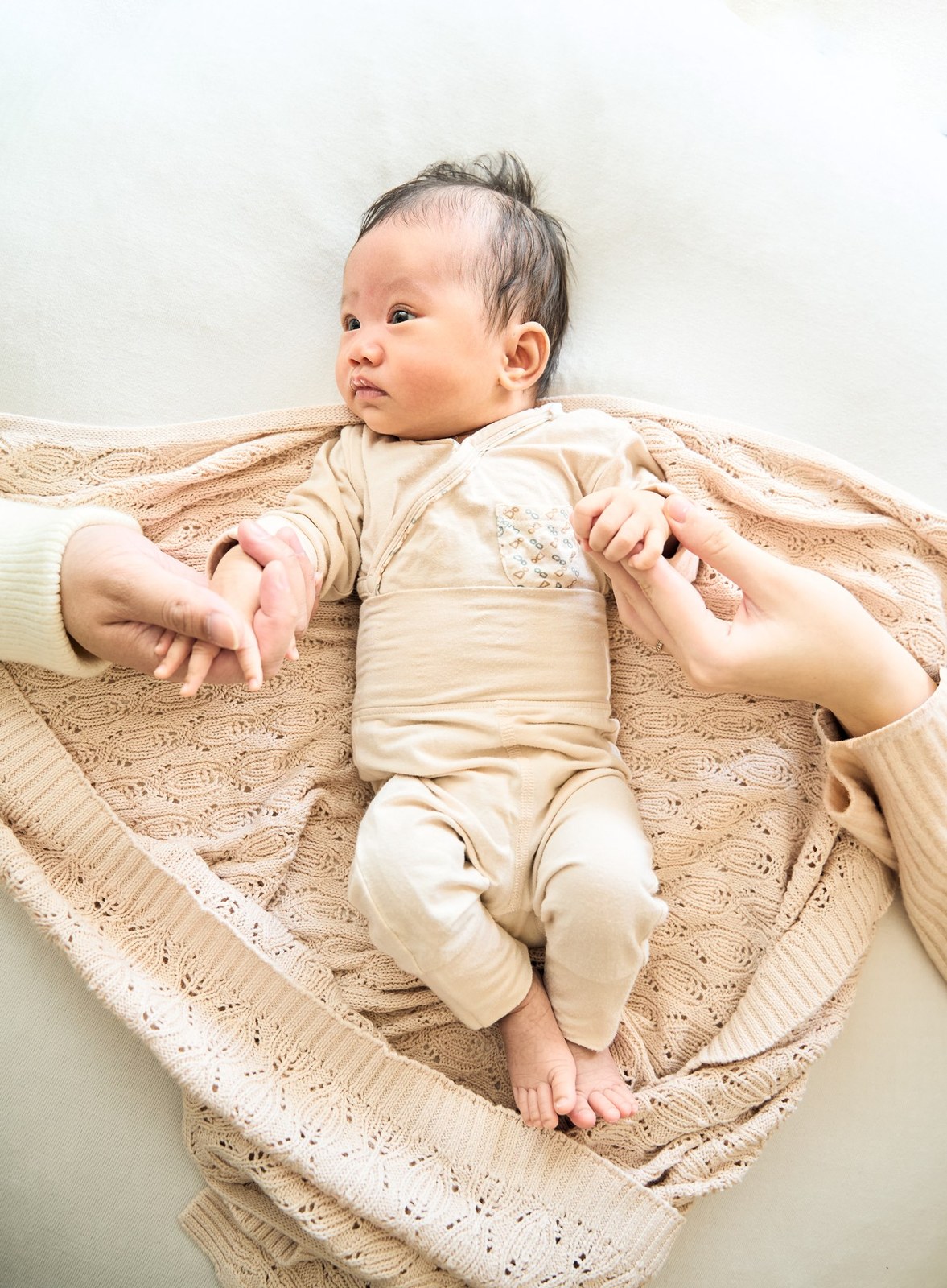 Newborn on a knitted blanket with her parents' hands either side