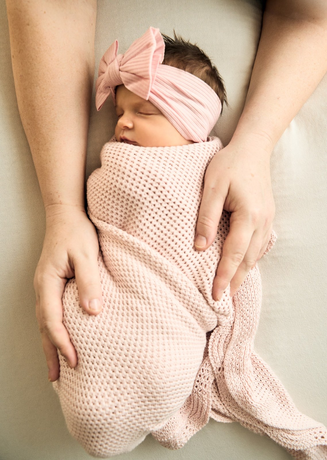 Swaddled newborn in a pink bow headband with her mum's hands resting nearby