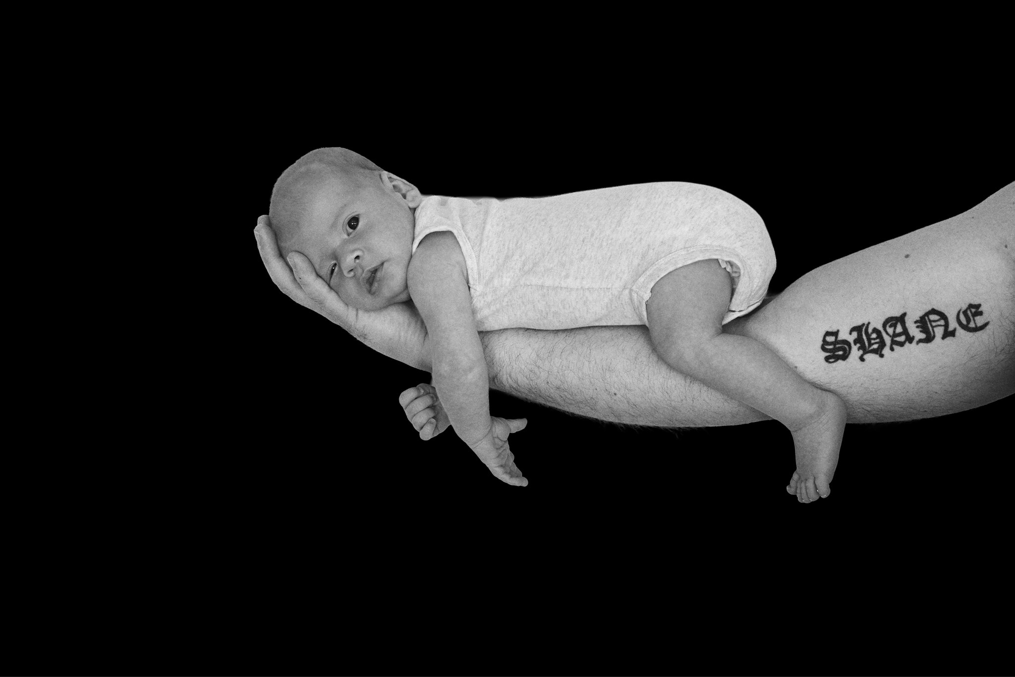 A newborn baby lying awake along his dad's tattooed forearm, black and white in-home newborn portrait