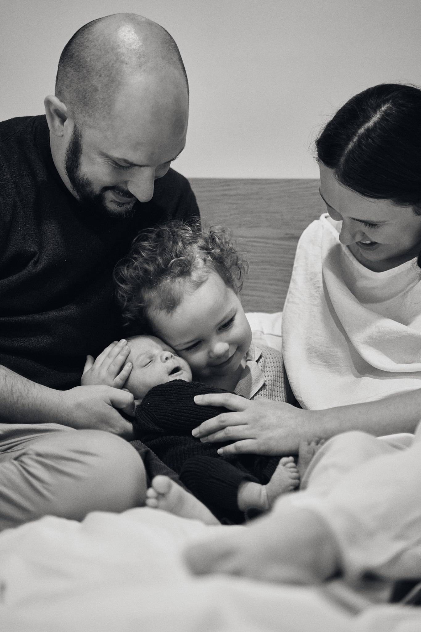 A curly-haired toddler cradling his newborn sibling while his parents look on, black and white in-home family session