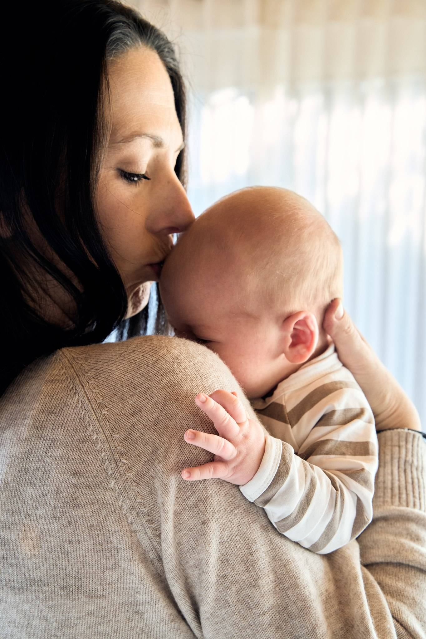 A mum kissing her newborn over her shoulder by a bright window, in-home newborn session