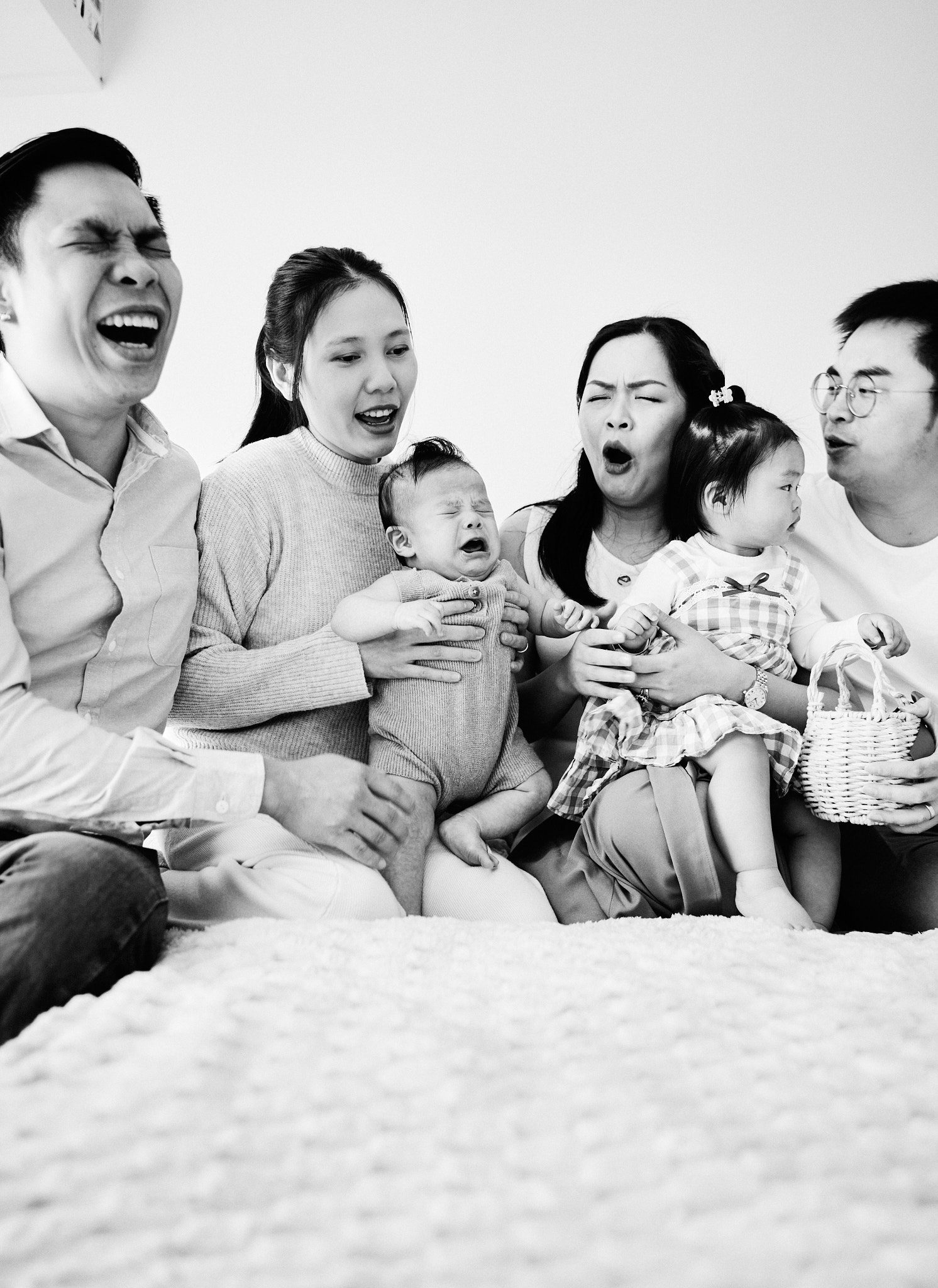 A family laughing and pulling faces together on the bed while the baby cries, black-and-white in-home session in South-East Melbourne
